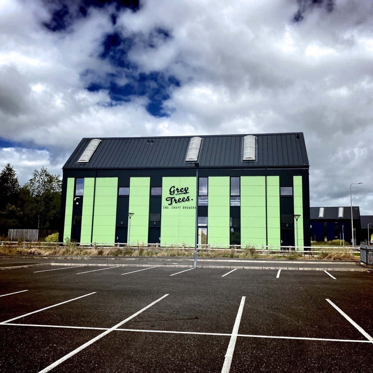 Grey Trees Brewery A modern building with a grey roof, green and grey walls, labeled Grey Trees, stands adjacent to an empty parking lot under a partly cloudy sky.