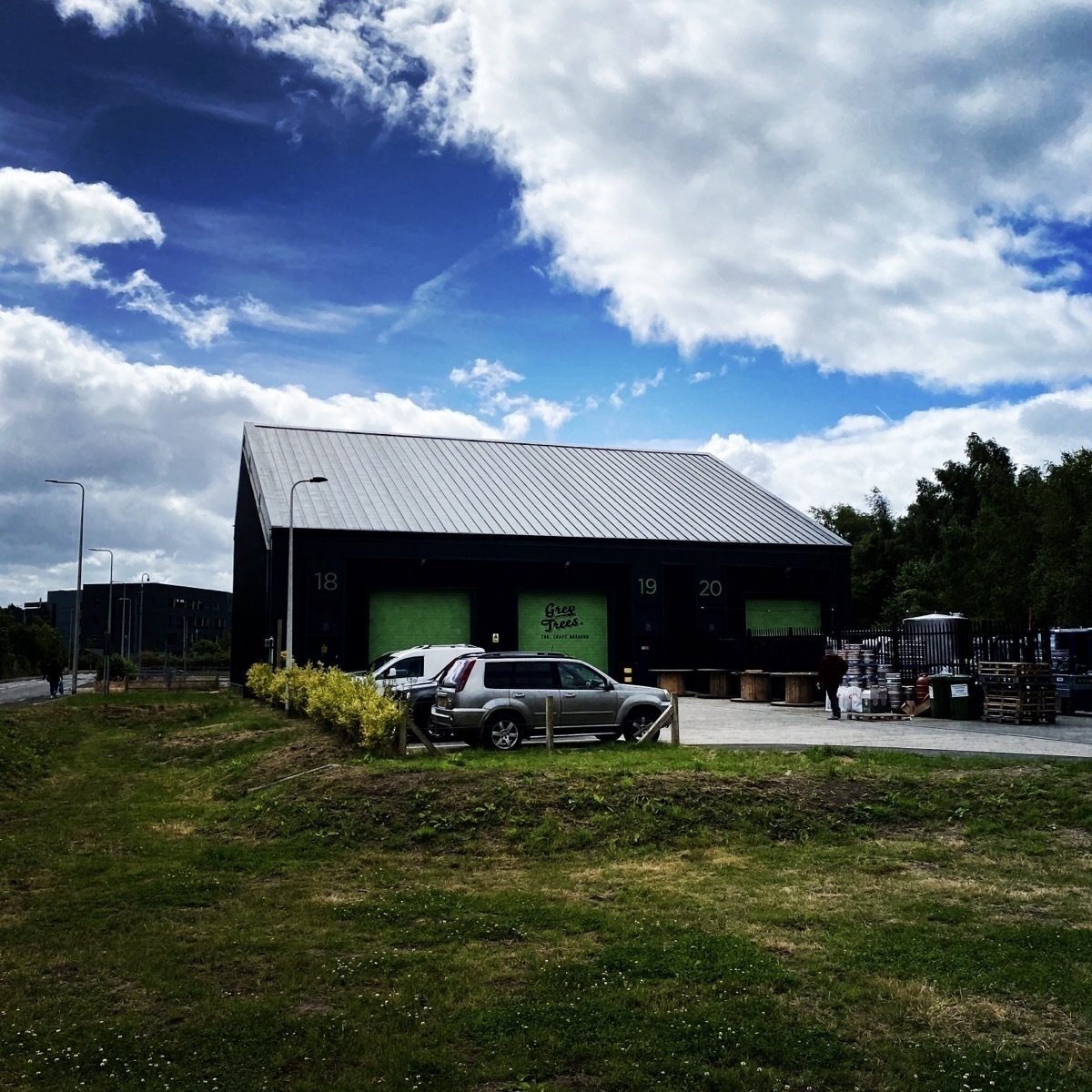 Grey Trees Brewery A dark-colored warehouse with the numbers 18, 19, and 20 on its doors is surrounded by parked cars and greenery under a partly cloudy sky.