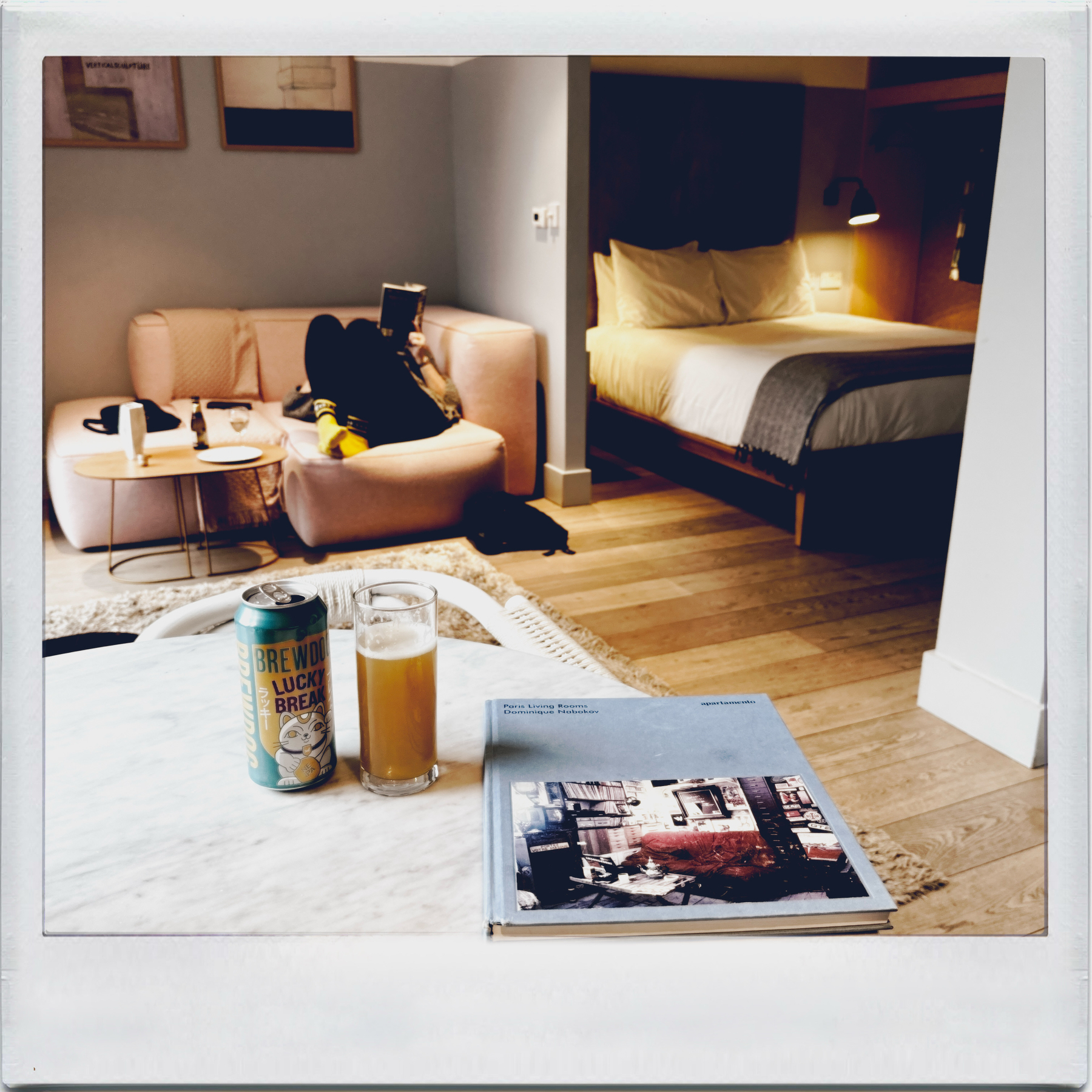 Auto-generated description: A cozy living space is depicted with a person relaxing on a couch, a can and glass of beer on the table, and a book beside them.