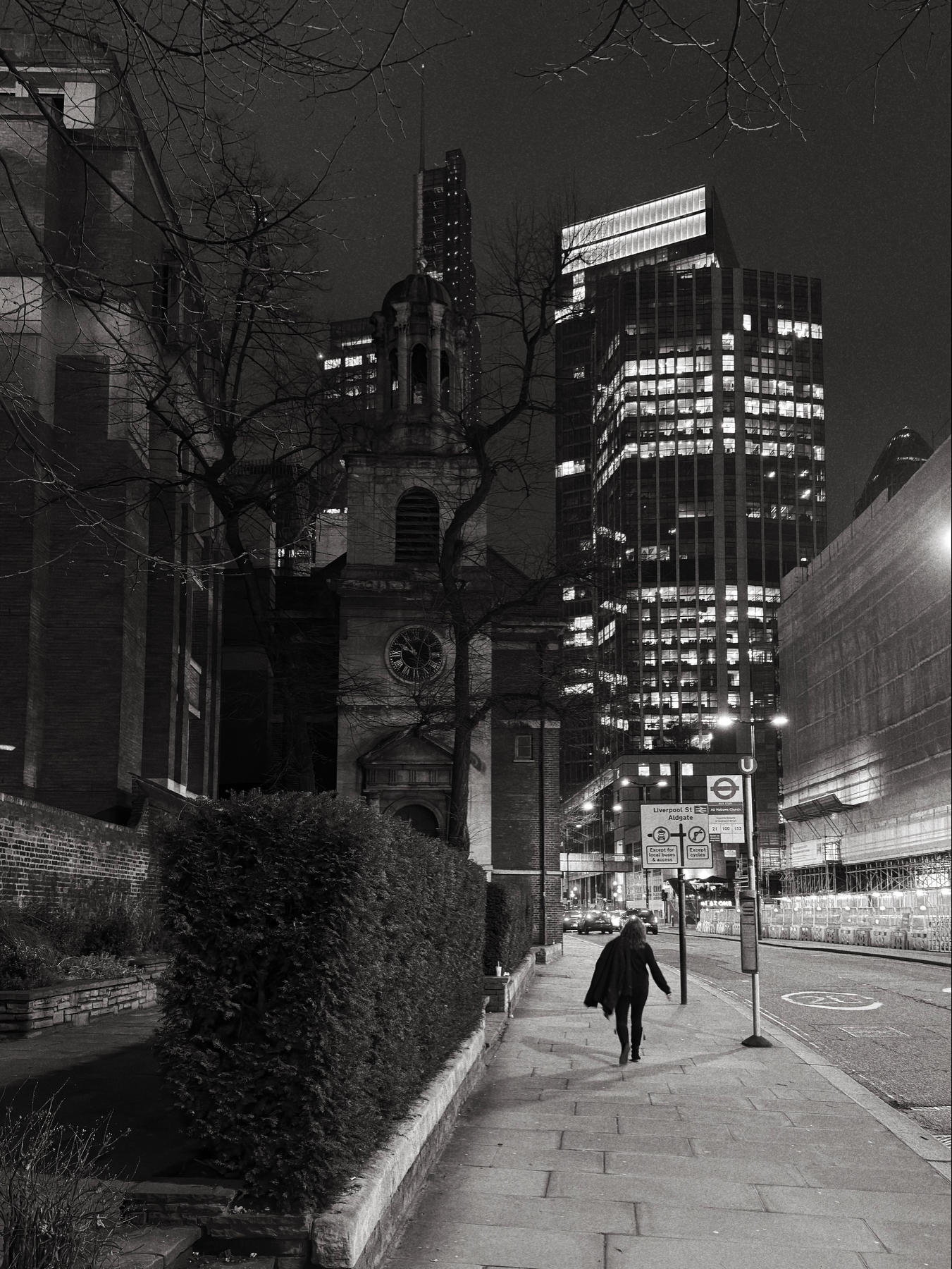 Auto-generated description: A solitary figure walks along a city street at night, framed by towering buildings and an old church.