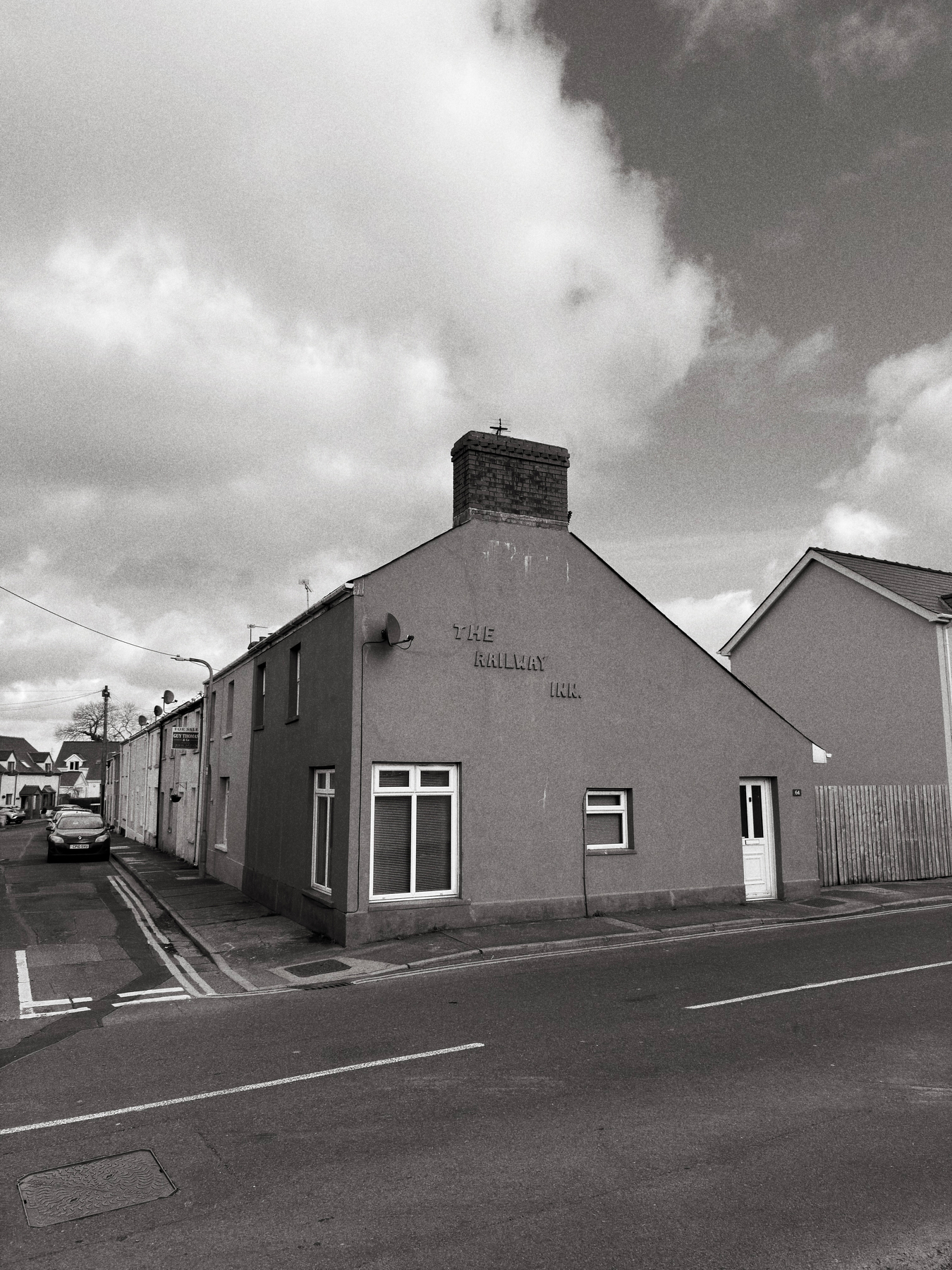 Auto-generated description: A road runs alongside a building marked The Railway Inn, with several other structures and parked cars in the background under a cloudy sky.