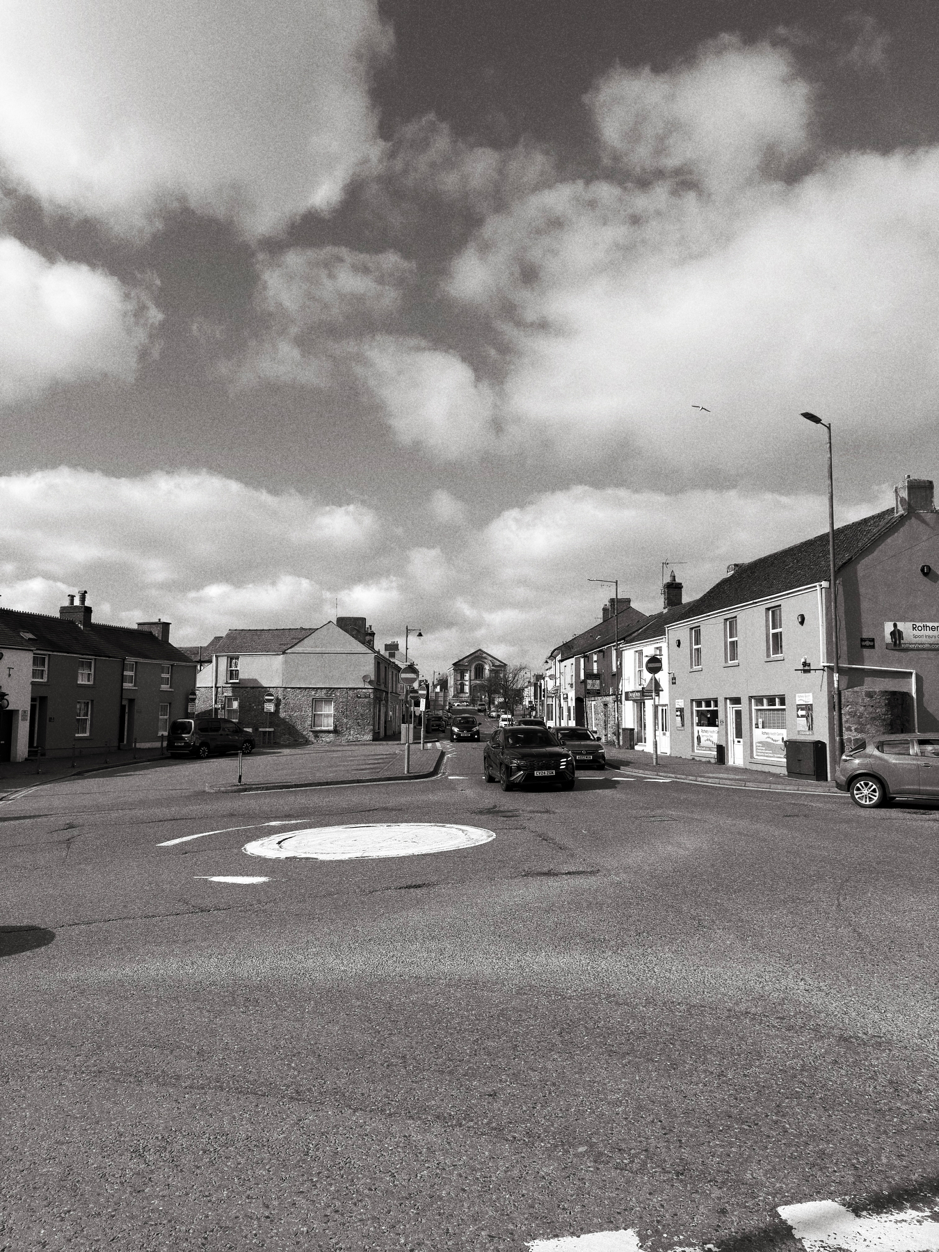 Auto-generated description: A monochrome street scene shows a roundabout in a small town with vehicles, buildings, and cloudy skies overhead.
