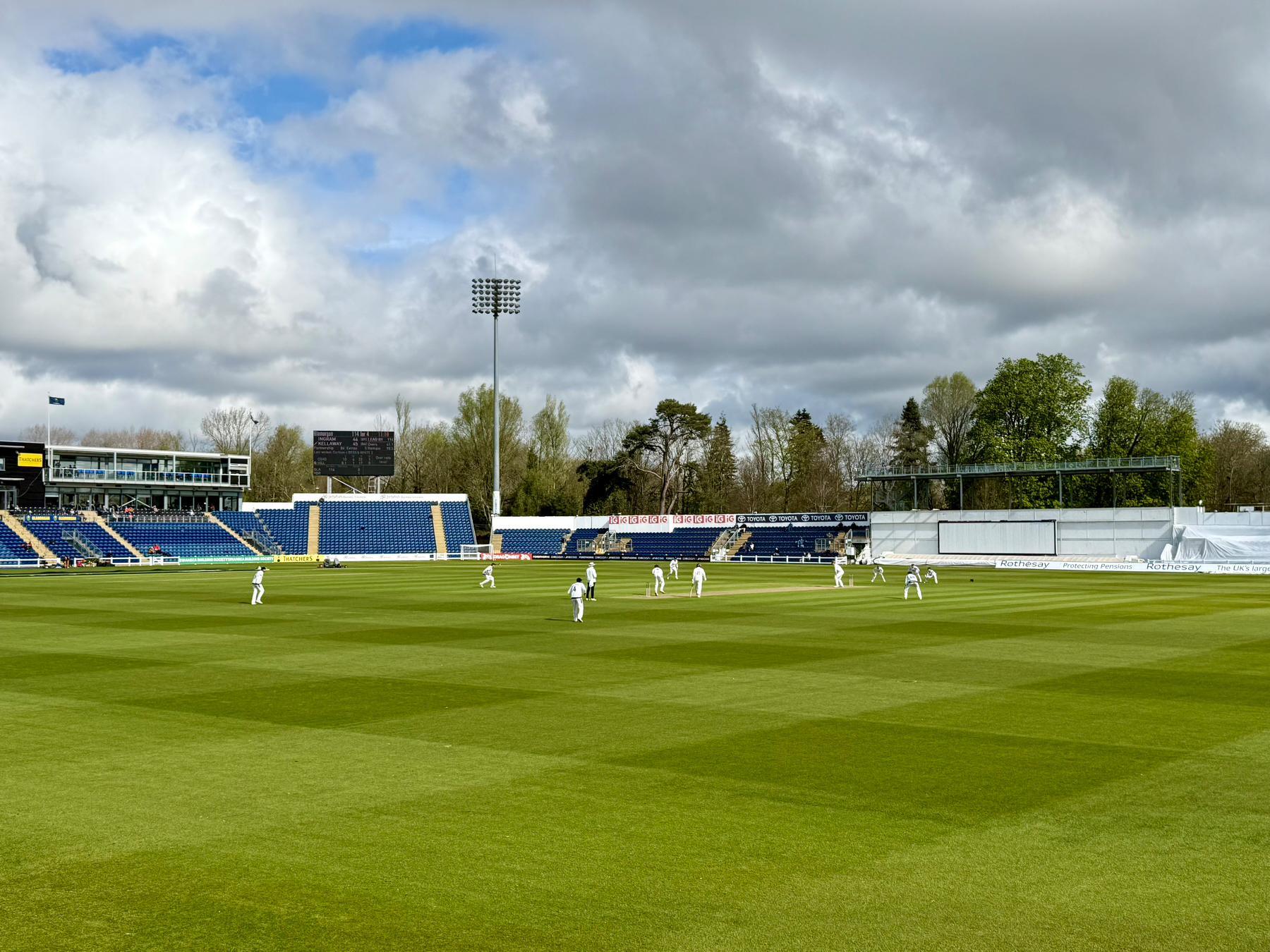 A cricket match is taking place on a green field under cloudy skies, surrounded by stadium seating.