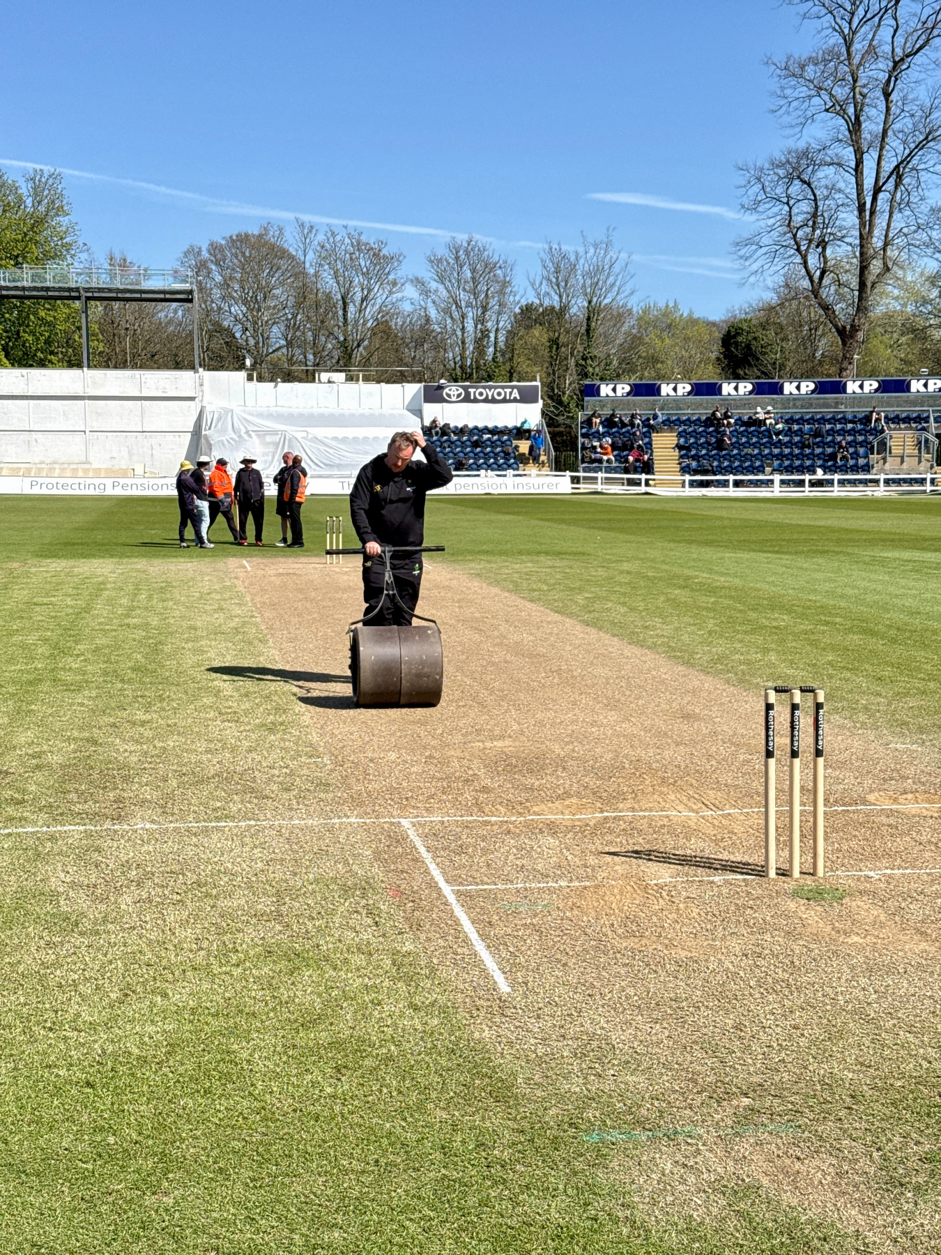 Auto-generated description: A person is operating a roller machine on the cricket pitch of a stadium under a clear sky.