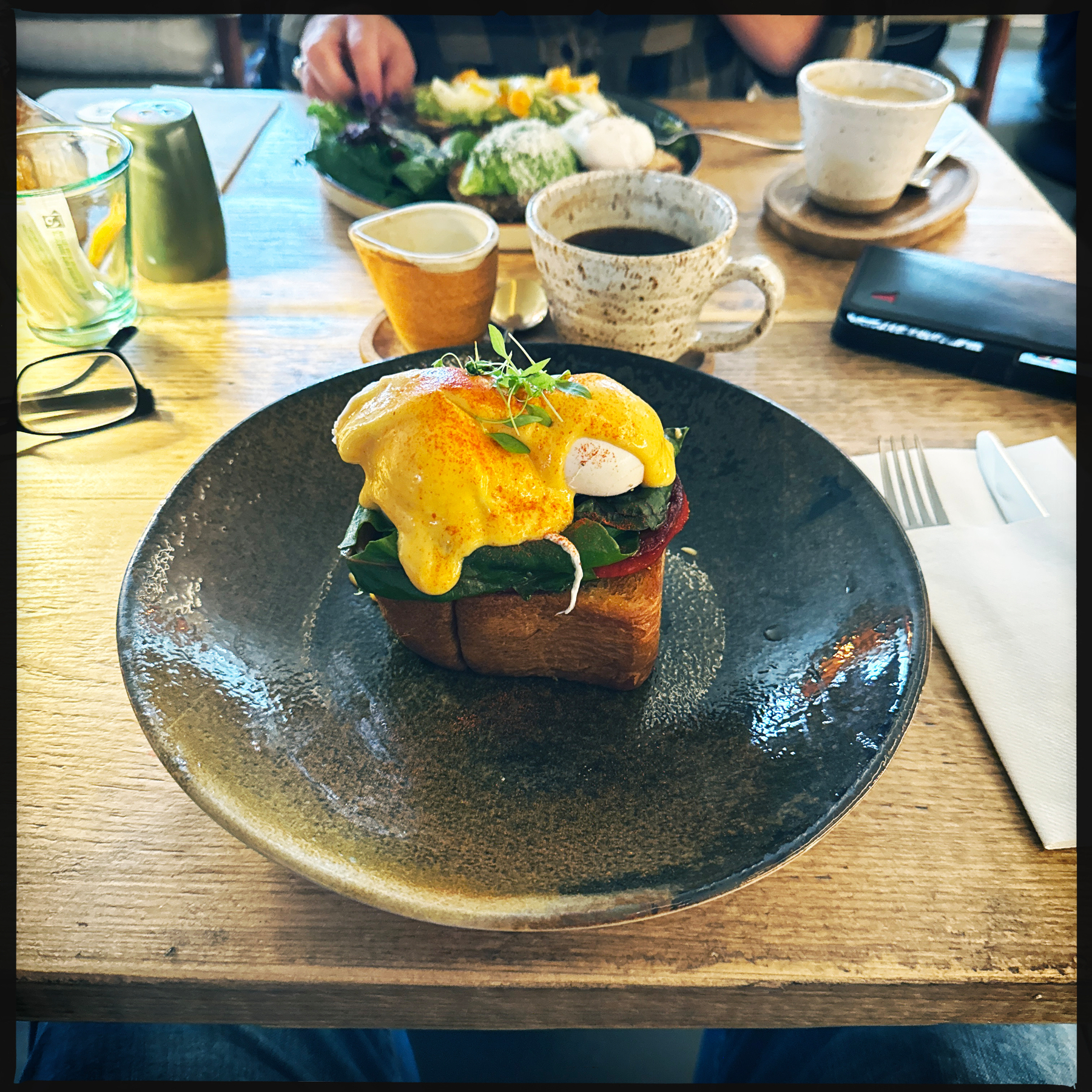 A delicious eggs Benedict dish with hollandaise sauce is served on toast, placed on a rustic plate, accompanied by various cups and utensils on a wooden table.