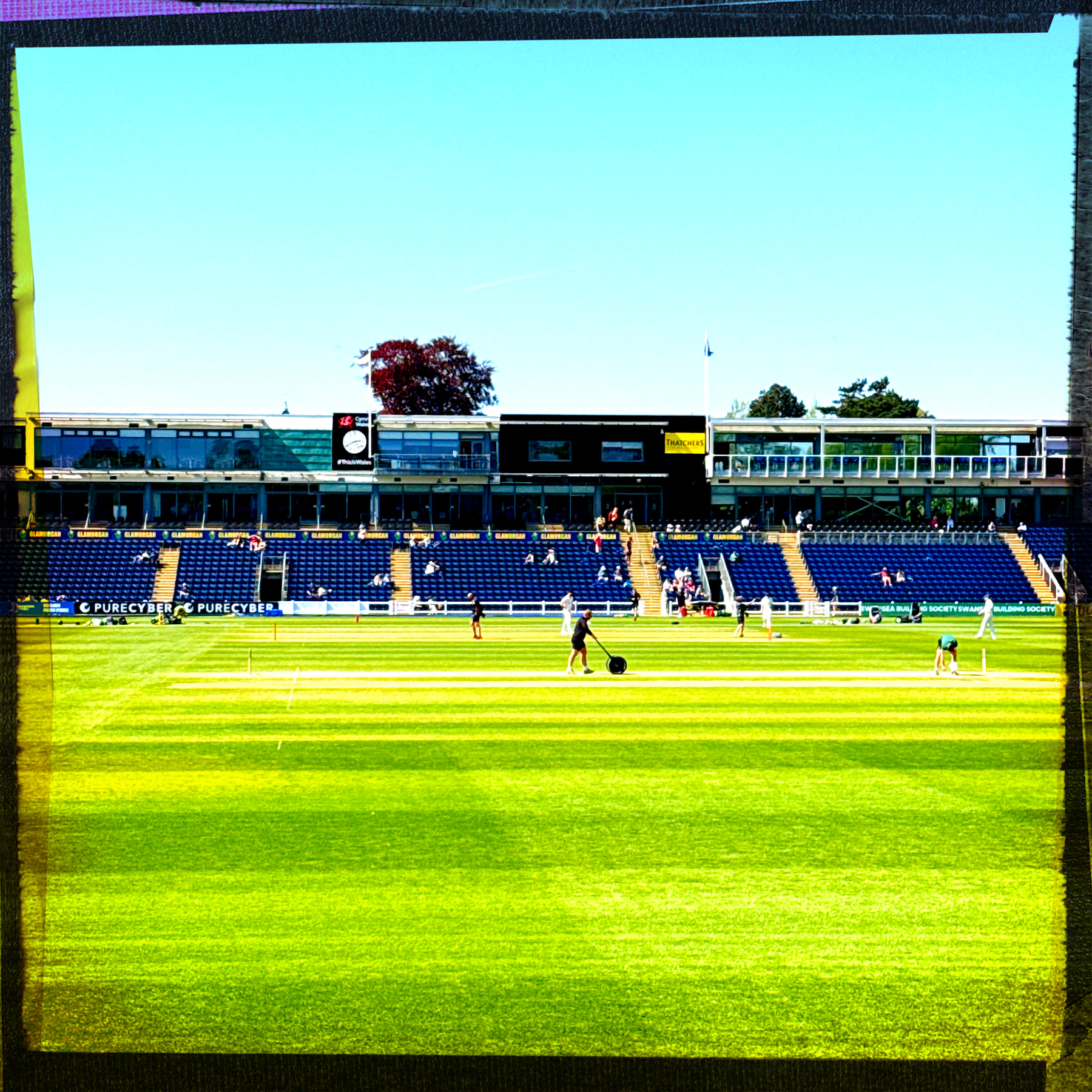 A cricket field features people preparing the pitch with empty stands and a two-story pavilion in the background.