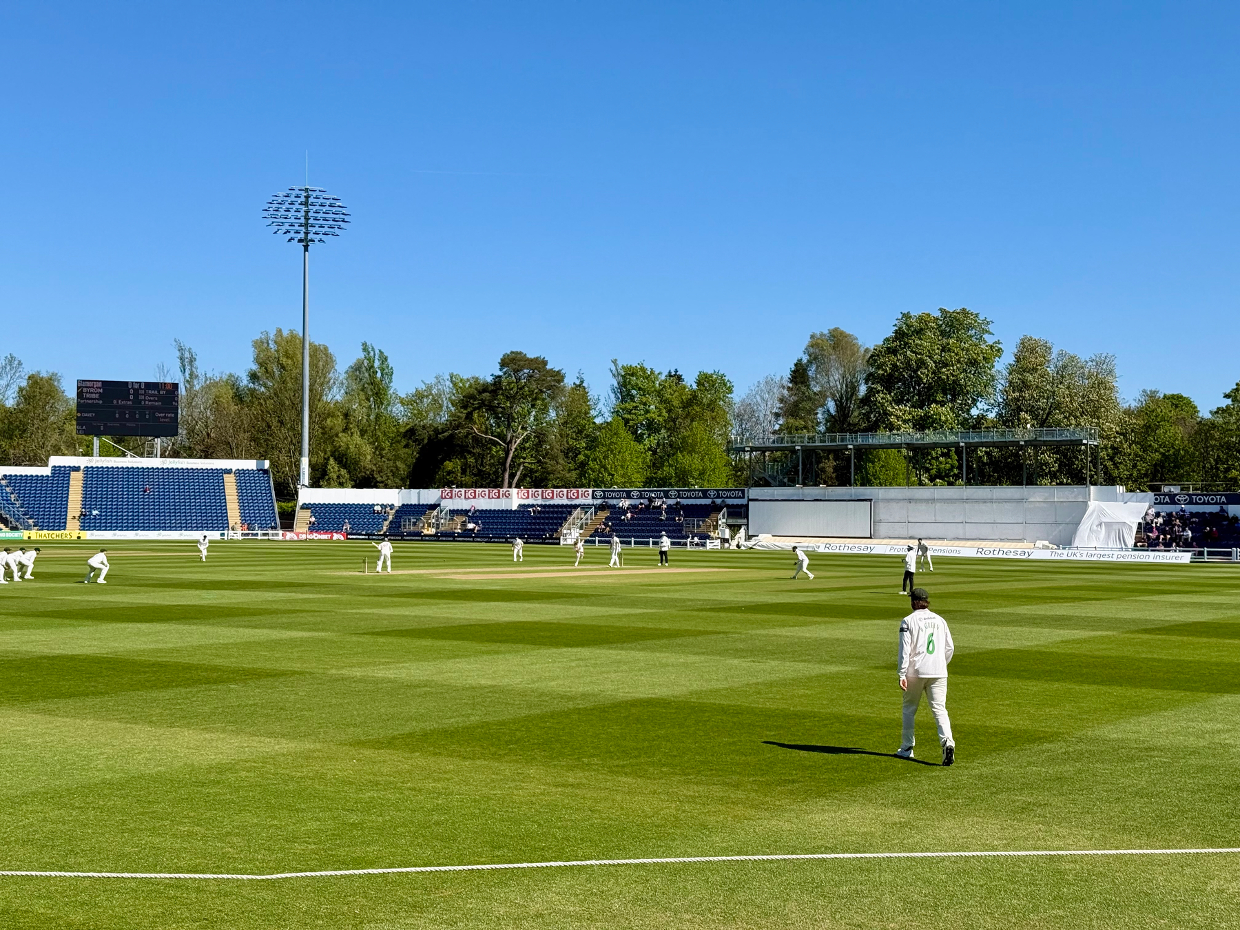 A cricket match is taking place on a sunny day in a stadium with players on the field and spectators in the stands.