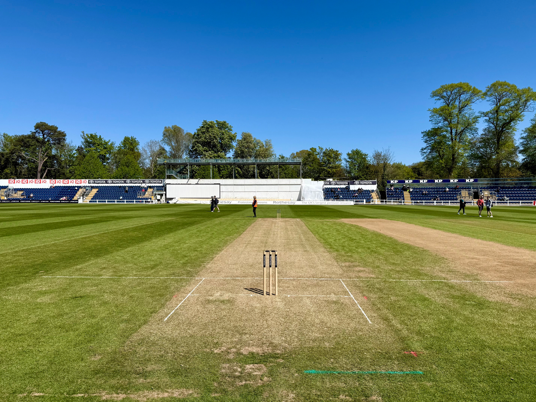 A cricket pitch set in an outdoor stadium with clear blue skies and players in the background.