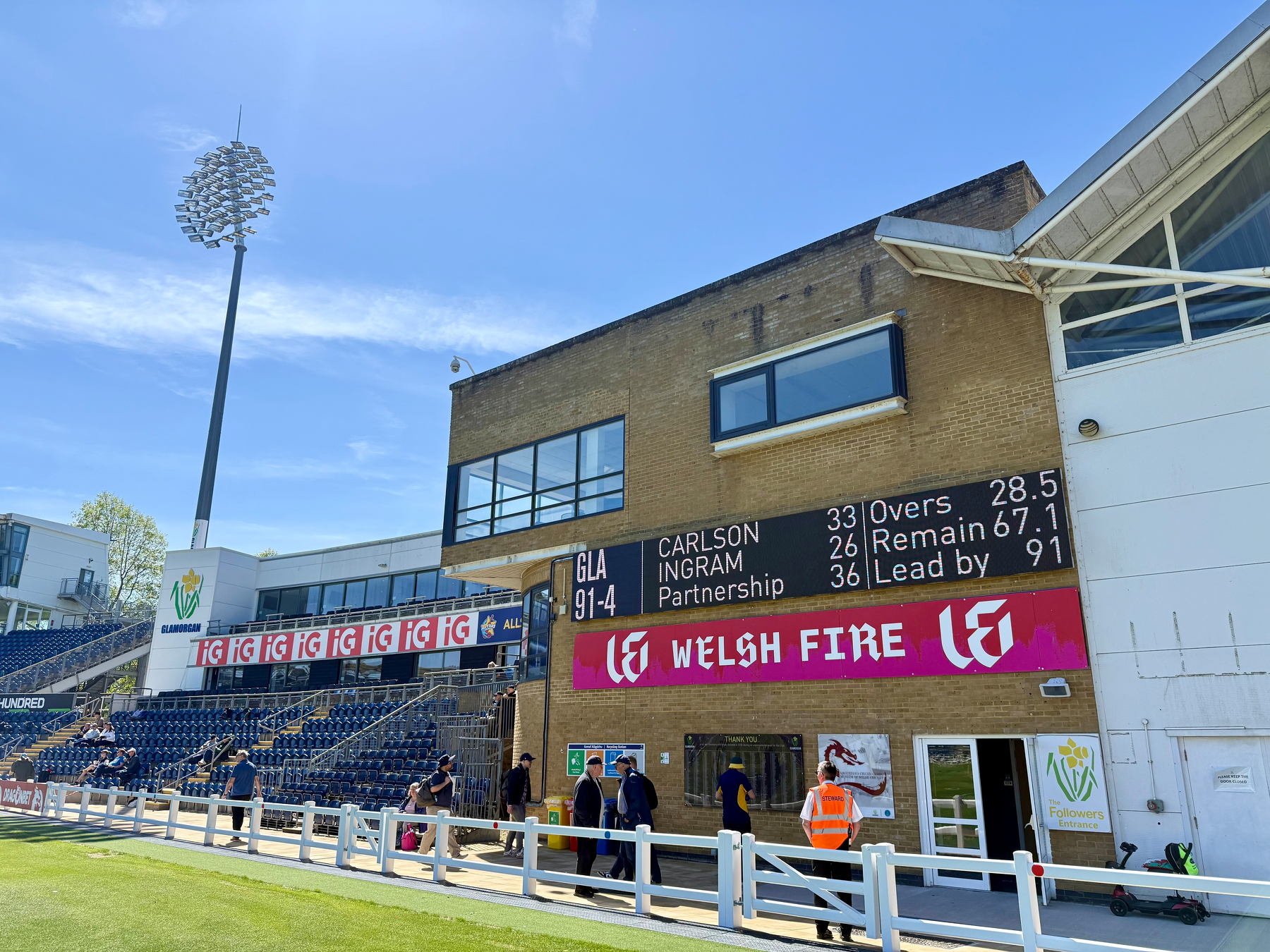 A cricket scoreboard is displayed at a stadium, showing details for a match involving Welsh Fire, with people and seating visible in the background.
