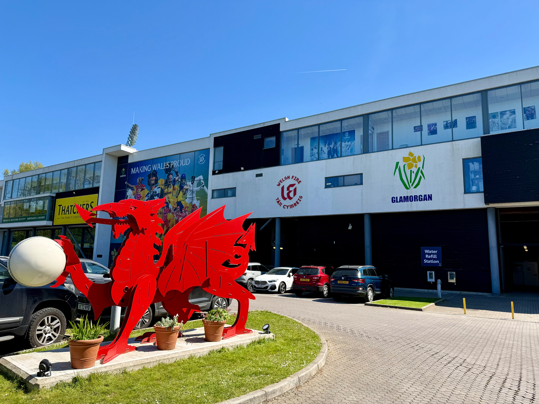 A red dragon sculpture stands outside a sports building with signs for Glamorgan, amidst parked cars and greenery.