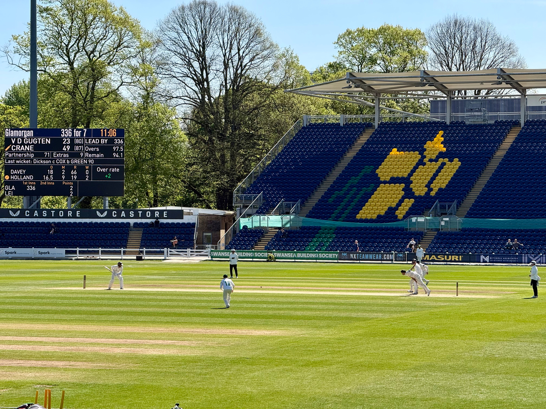 A cricket match is taking place in a stadium with a scoreboard displaying scores and a stand featuring a yellow emblem on blue seats.