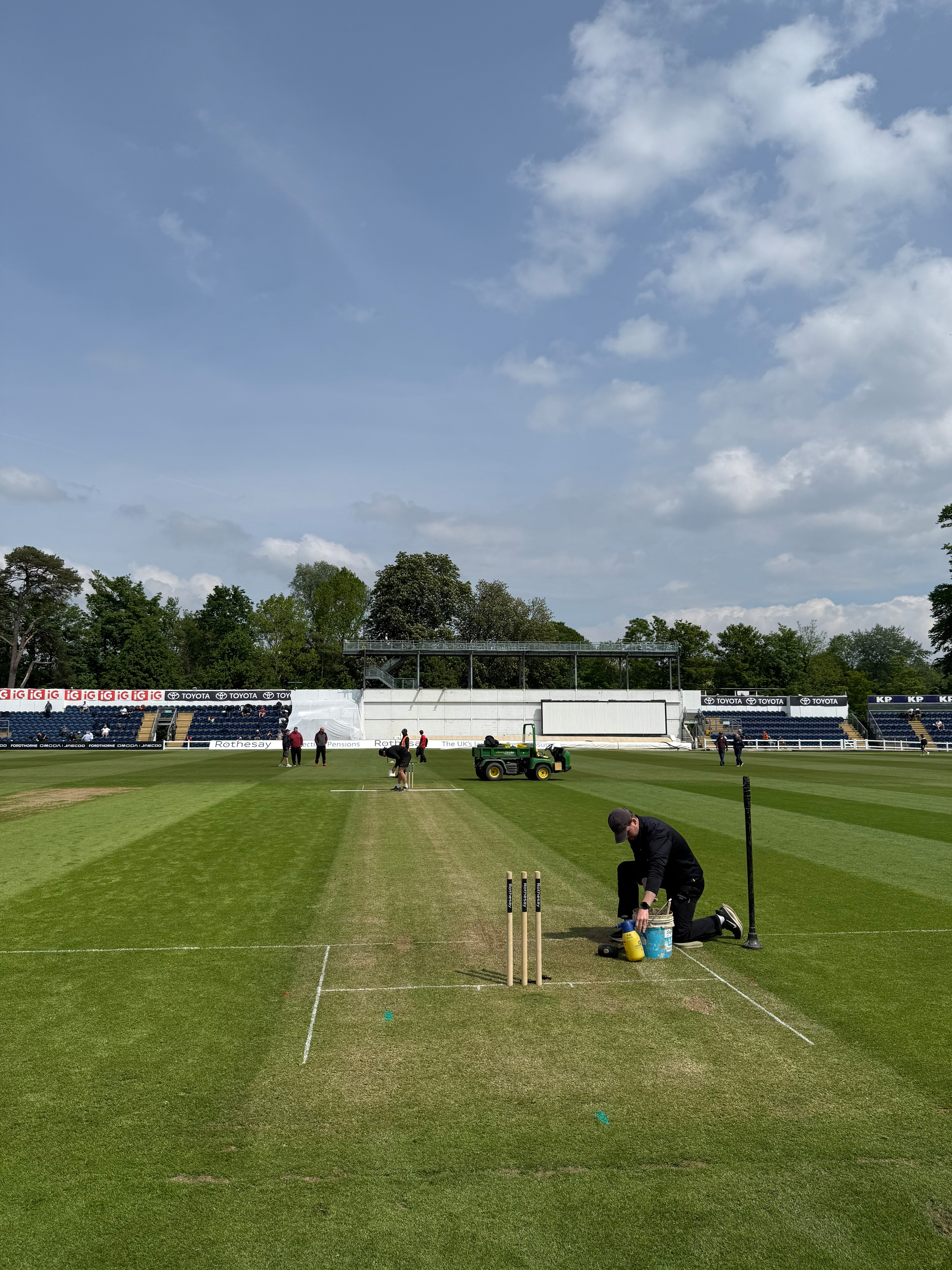 Auto-generated description: A groundskeeper is preparing a cricket pitch on a sunny day, with seating and lush trees in the background.