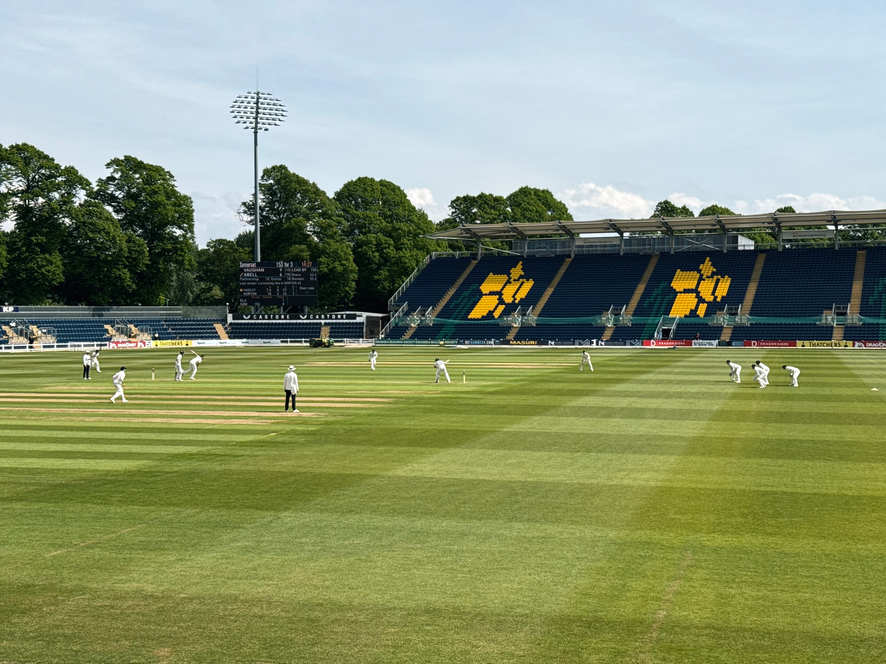 A cricket match is taking place on a large, lush green field with players in white uniforms and a mostly empty stadium in the background.