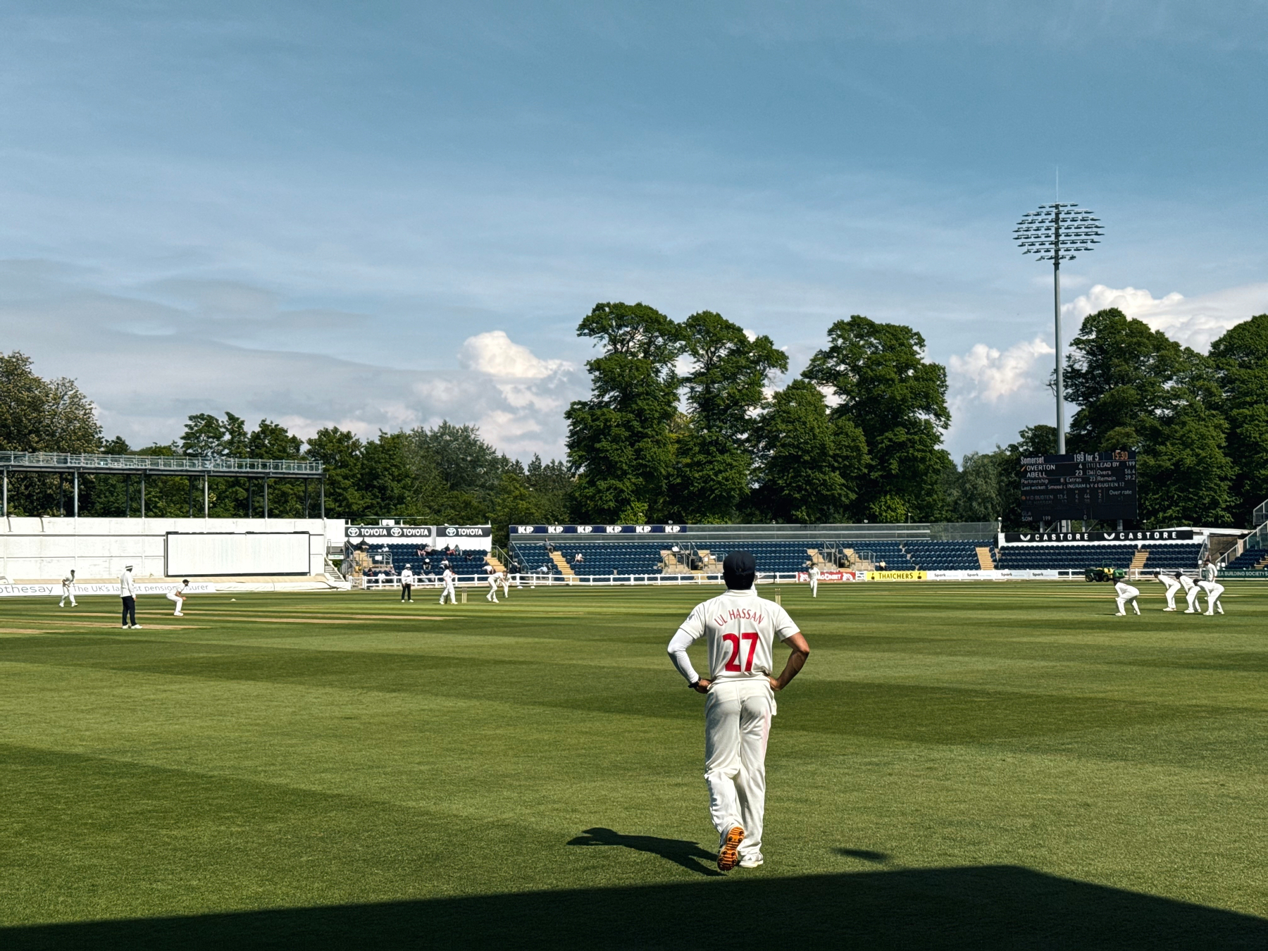 A cricket match is taking place on a sunny day with a player wearing the number 27 on the field.