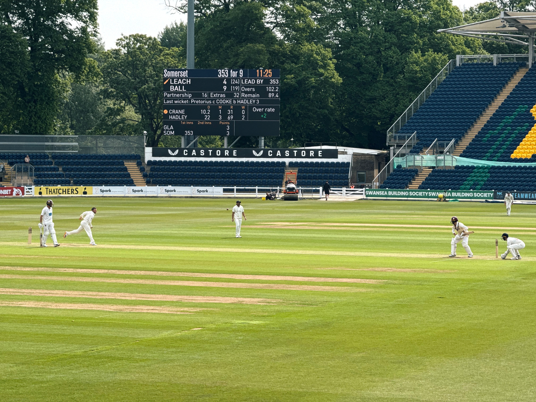 A cricket match is in progress on a grassy field with players, umpires, and a scoreboard in the background.