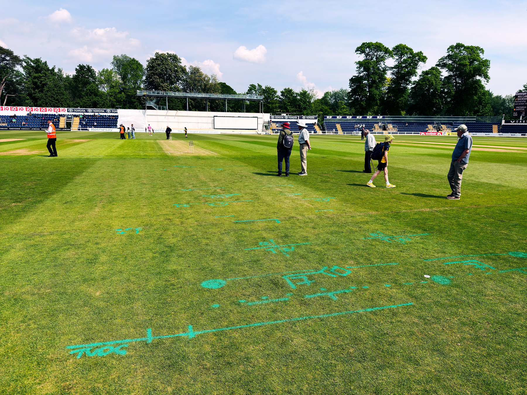 People are walking across a cricket field with marked diagrams and symbols on the grass.
