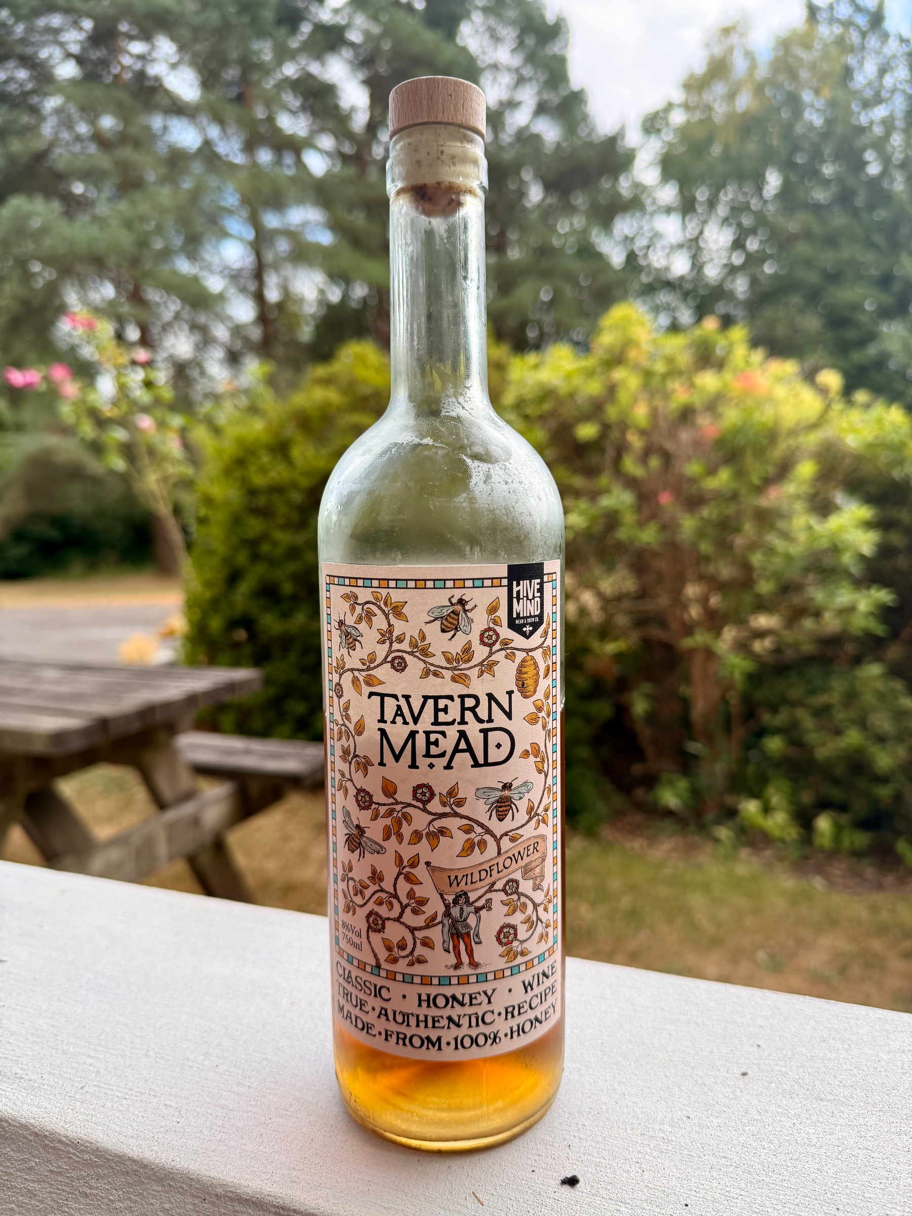 A bottle of Tavern Mead Wildflower honey wine, partially filled and sealed with a cork, is placed on a white surface with a garden and wooden picnic table in the background.