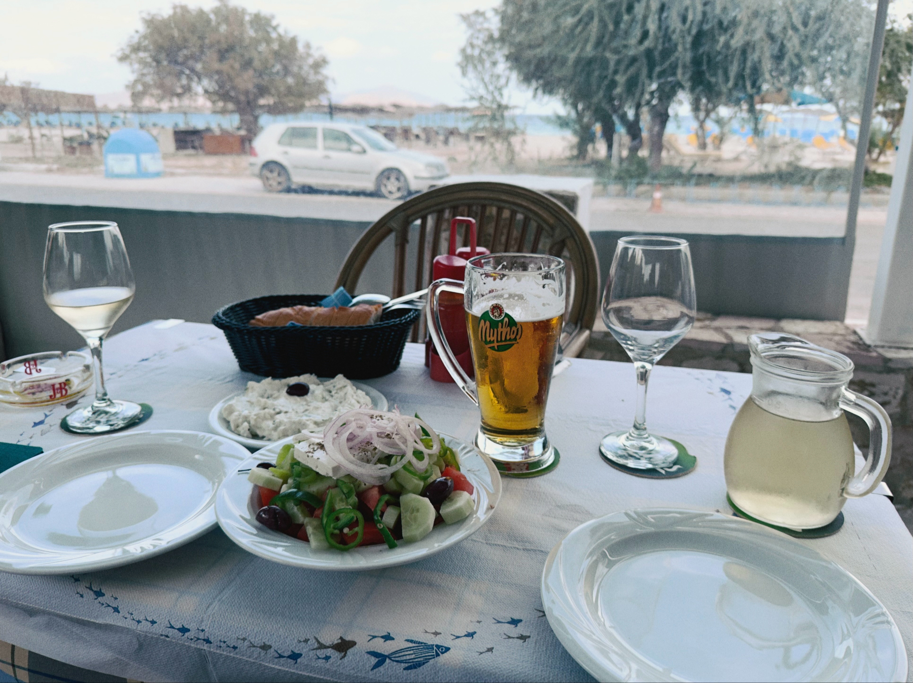 A table is set with plates, glasses of wine and beer, a salad, bread, and a carafe, with a scenic view of trees, cars, and a body of water in the background.