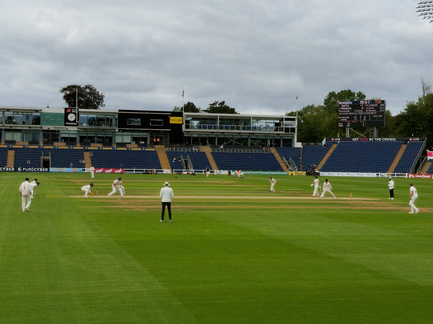 A cricket match is in progress on a green field with players positioned and a scoreboard visible in the background along with spectators' seating.