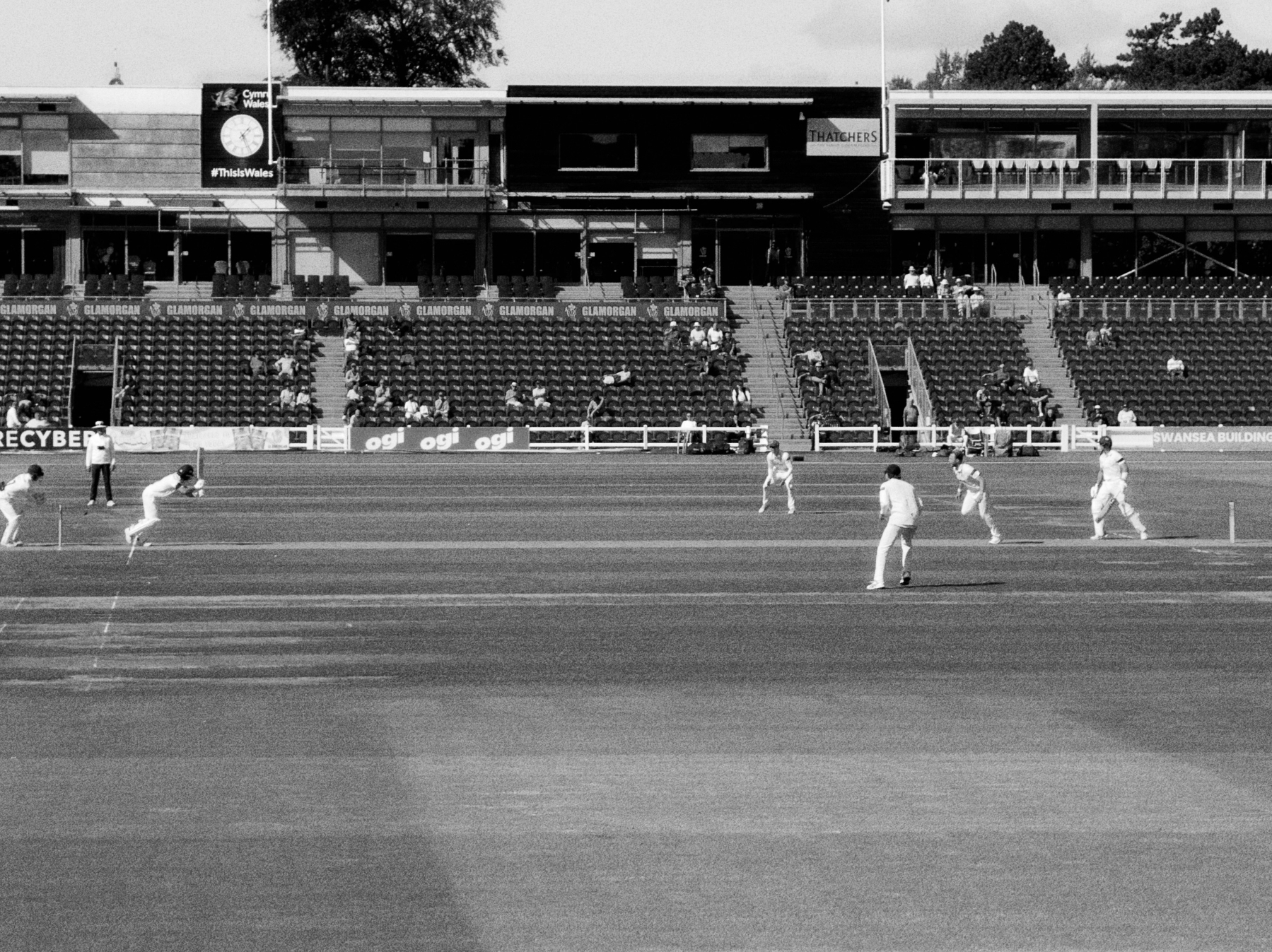 A cricket match is taking place on a field with players positioned and a nearly empty stadium in the background.